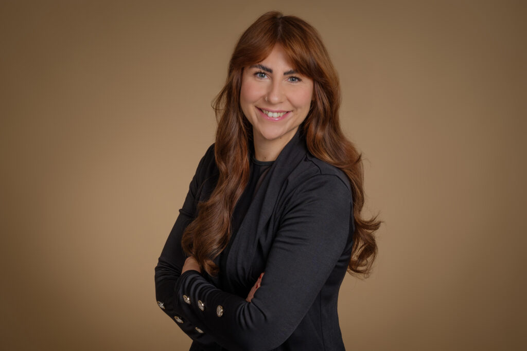 Frau mit roten Haaren lächelt, schwarzer Blazer, brauner Hintergrund.